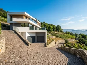 Nell'immagine si vede una moderna casa a più livelli, costruita su una collina particolarmente ripida. Un elegante viale d'accesso, lastricato con cubetti di porfido, arriva con una forte inclinazione sino a due porte sezionali per garage color antracite.