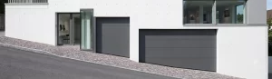 Nell'immagine si vede una strada in salita. Al lato della strada si vede una casa moderna bianca con due porte sezionali per garage, color antracite, che si affacciano direttamente sul marciapiede della strada. Gli ingressi si trovano su una porzione di strada particolarmente inclinata. Per questo motivo gli ultimi pannelli inferiori delle porte del garage non hanno la classica forma rettangolare, bensì a trapezio, per seguire in modo complanare l'inclinazione della strada.