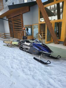 La foto mostra una motoslitta blu parcheggiata fuori dall'ingresso dell'hotel dove sono in corso i lavori. Sul retro si vede il carrellino della motoslitta carico di materiali utili per il montaggio delle porte automatiche.